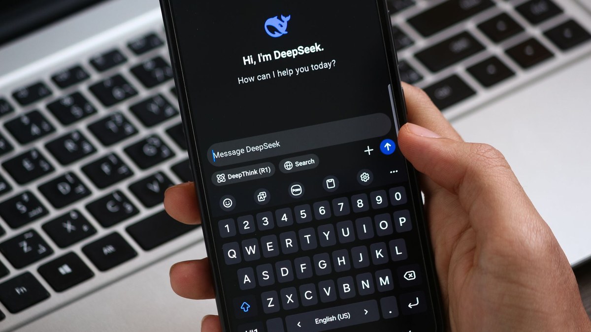 A hand holding a smartphone displaying the DeepSeek AI chat interface with an open keyboard in front of a laptop.