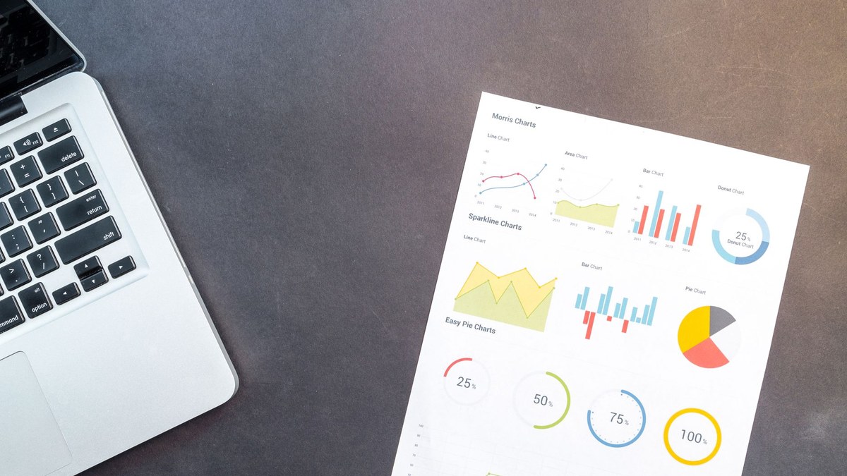 A silver laptop next to a printed sheet of paper displaying colorful data visualizations and charts on a dark grey desktop.